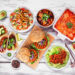 An image of a healthy plant based meal table scene. Top view on a white wood background. Jackfruit tacos, zucchini lasagna, walnut bolognese zoodles, chickpea burgers, hummus, soup, and chickpea salad.